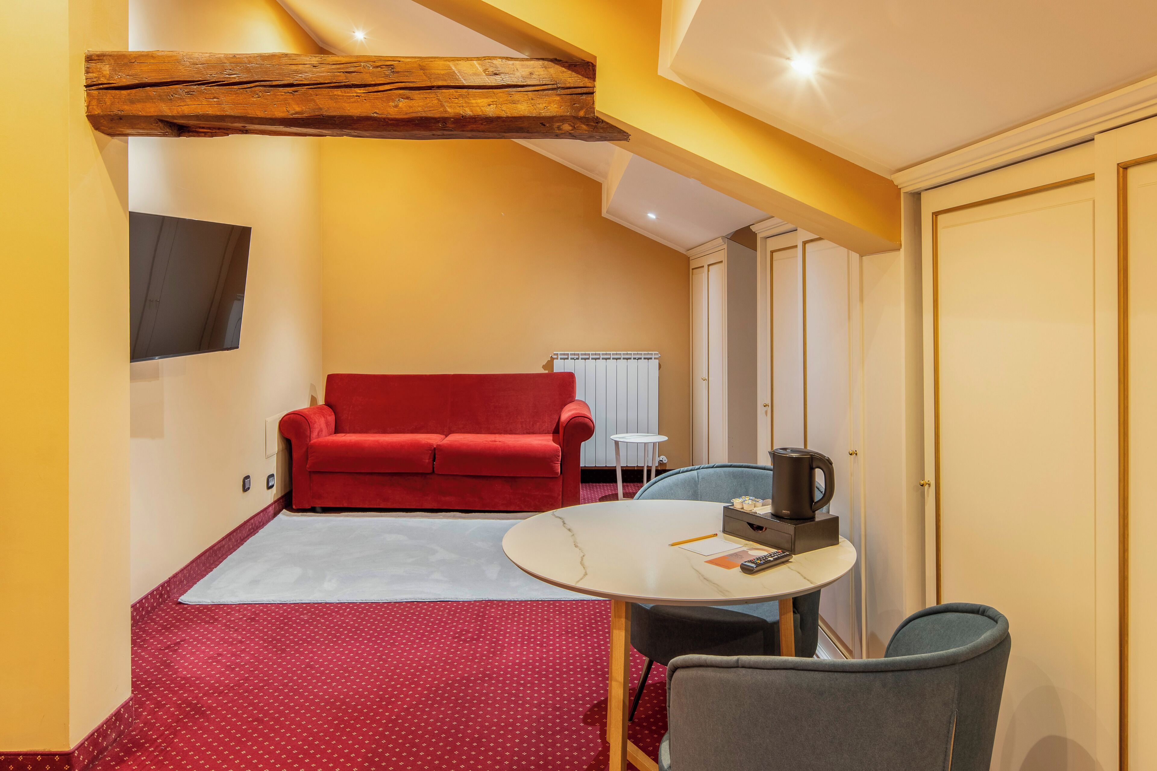 A compact living space with a red sofa, a small round table with a coffee maker, and a TV mounted on the wall.
