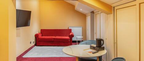 A compact living space with a red sofa, a small round table with a coffee maker, and a TV mounted on the wall.