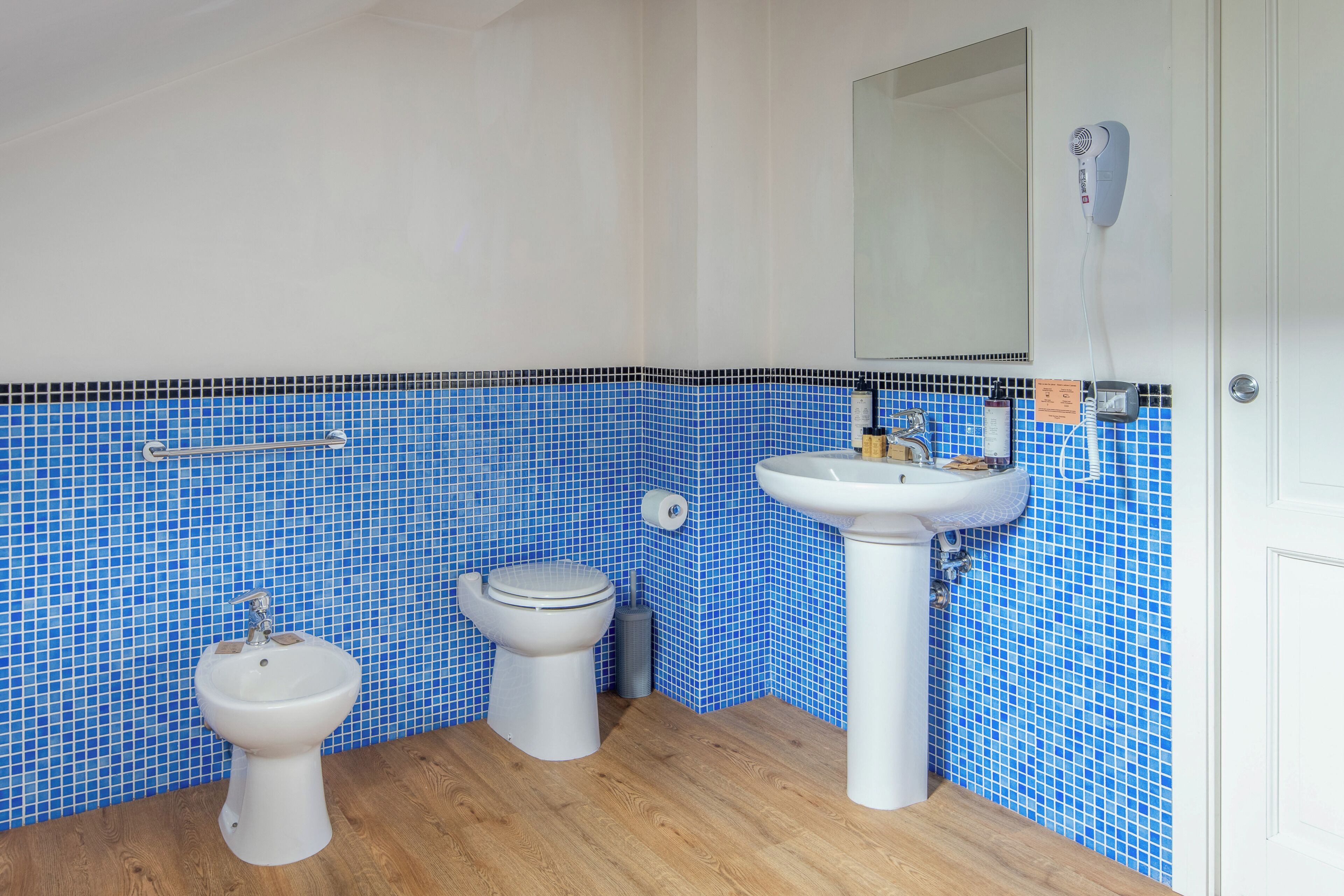 Un baño con inodoro, bidé y lavamanos, todos con accesorios blancos. Las paredes están revestidas con mosaicos azules.