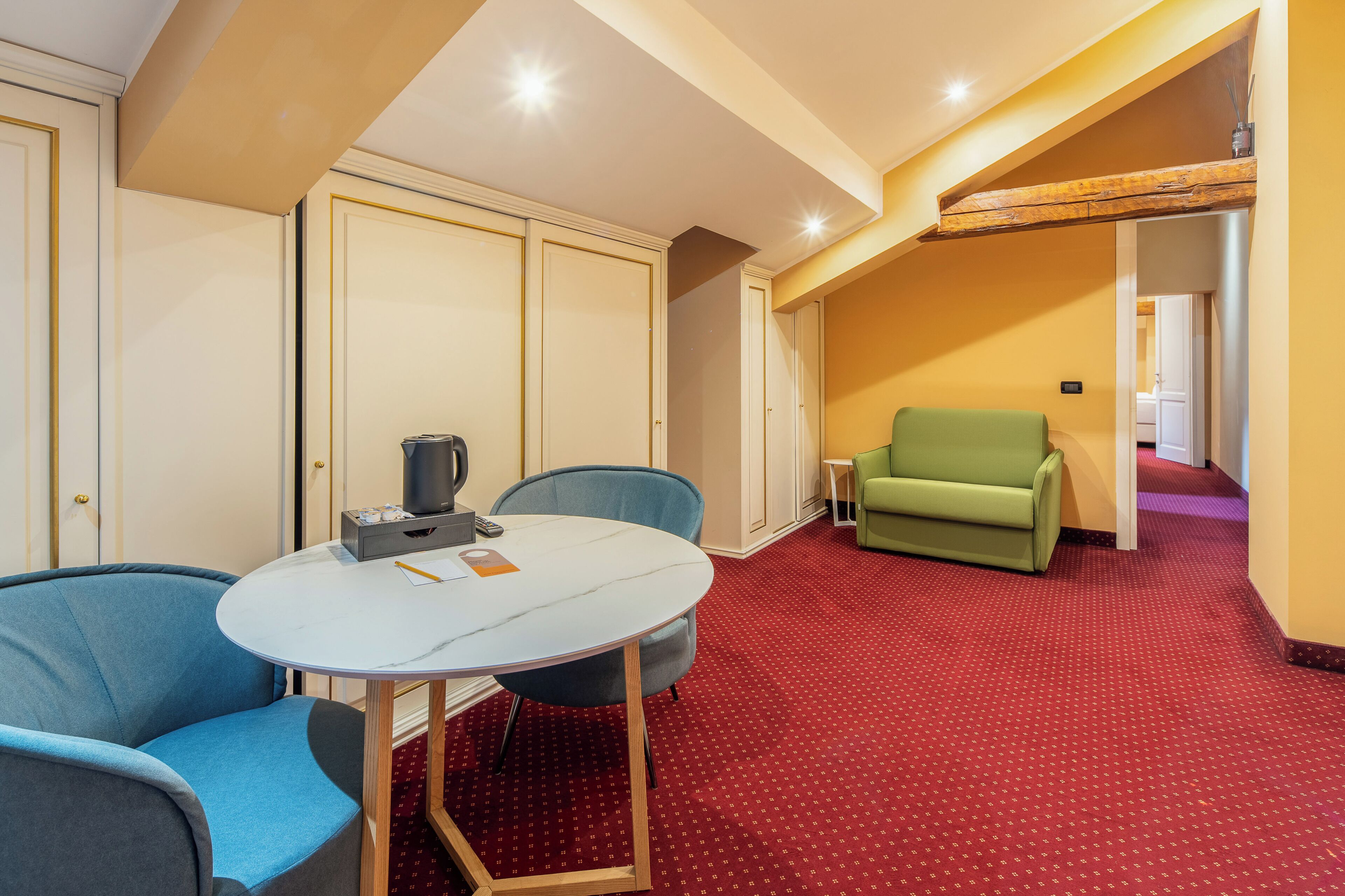 Habitació d'hotel amb una catifa vermella, una taula rodona amb cadires, un butac armchair verd i una nevera minibar.