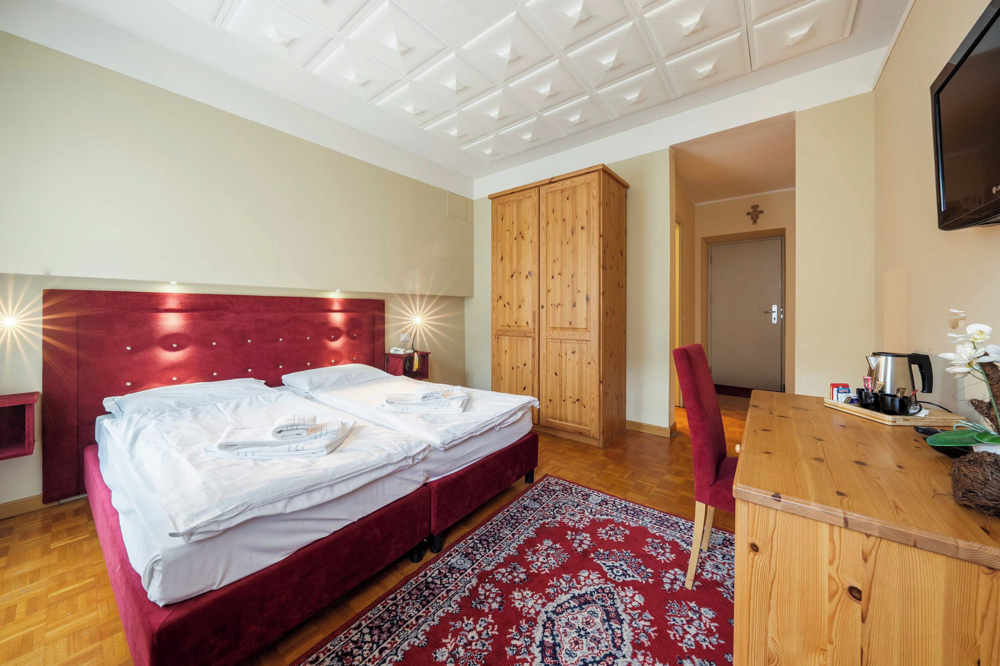 A bedroom with a red headboard, a wooden wardrobe, a desk with a chair, and a television on the wall.