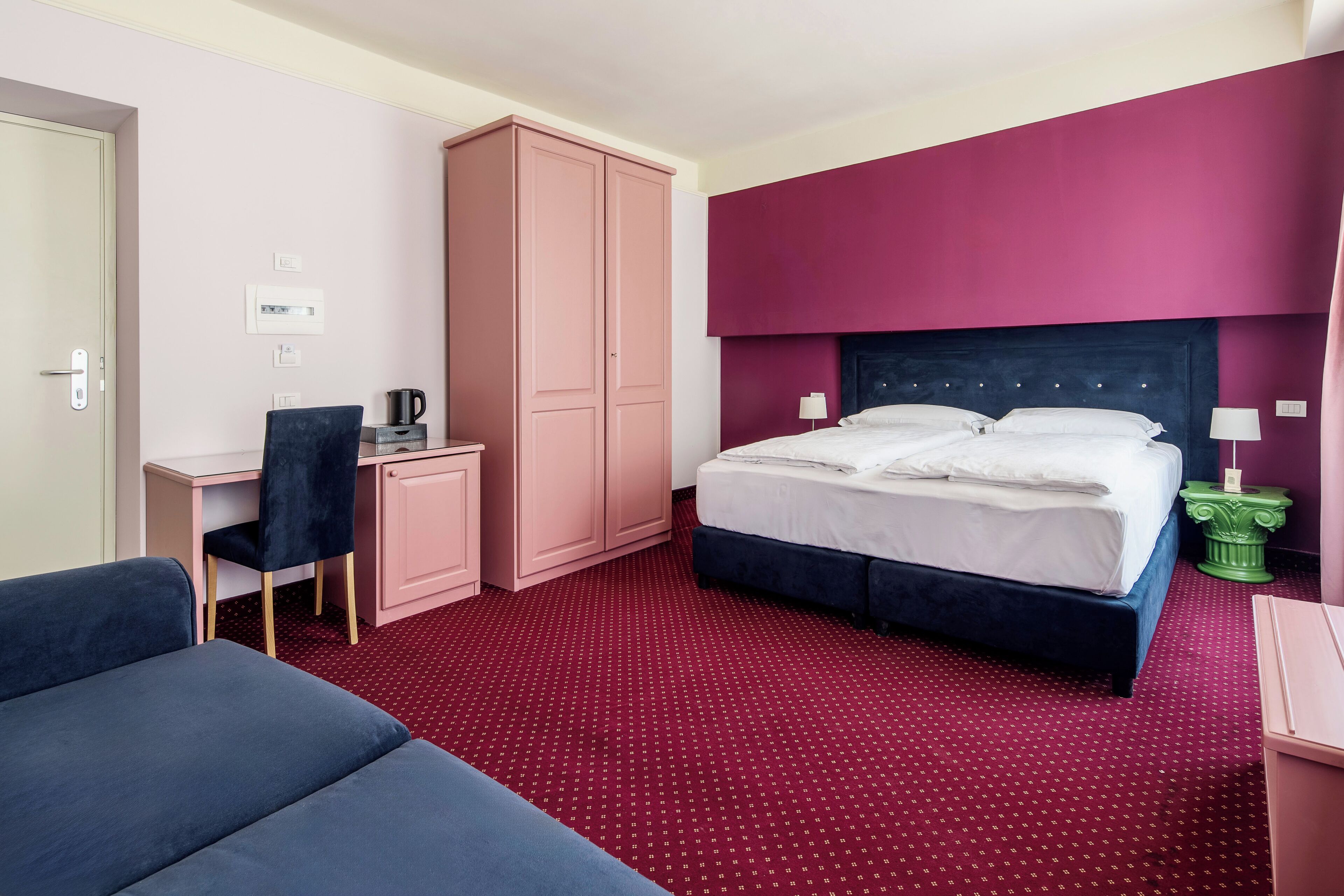 A hotel room with a bed, a desk, a chair, a wardrobe, and a red carpet.