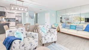A modern living room with a light blue wall, a beige sofa, two patterned armchairs, a dining area with a table and chairs, and a kitchen in the background. - Blue Dolphin (Santa Rosa Beach)