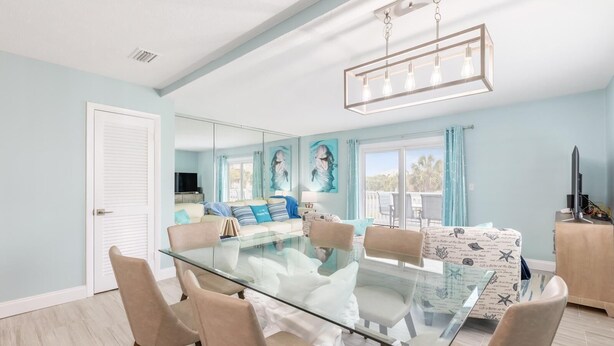 A modern living room with a glass dining table, beige chairs, a sofa with patterned cushions, and a large window with curtains. - Blue Dolphin (Santa Rosa Beach)