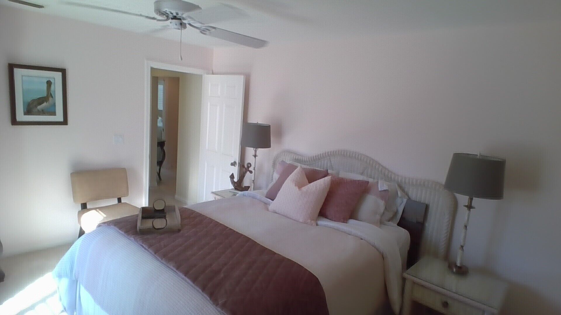 A bedroom with a bed, two bedside tables with lamps, a ceiling fan, and a door leading to another room.