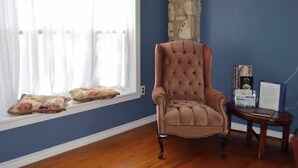Une pièce avec un mur bleu, un mur en pierres apparentes, une fenêtre avec des rideaux blancs, un fauteuil à capitons et une petite table en bois sur laquelle sont posés divers objets.