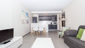 A modern living room with a flat-screen TV, a white coffee table, a gray sofa with a green pillow, and a colorful chair. There is a dining area with a white table and chairs, and a kitchen in the background. - 05 -b902 in Portim o (Portimão)