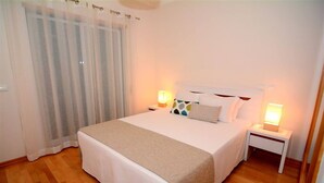 Ein ordentlich gemachtes Bett mit weißer und beiger Bettdecke, zwei Nachttische mit Lampen und ein Fenster mit durchsichtigen Vorhängen.