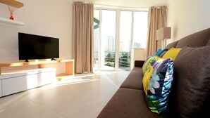 Ein modernes Wohnzimmer mit Flachbildfernseher, einer Couch mit bunten Kissen, einem kleinen Tisch und einem großen Fenster mit Vorhängen.