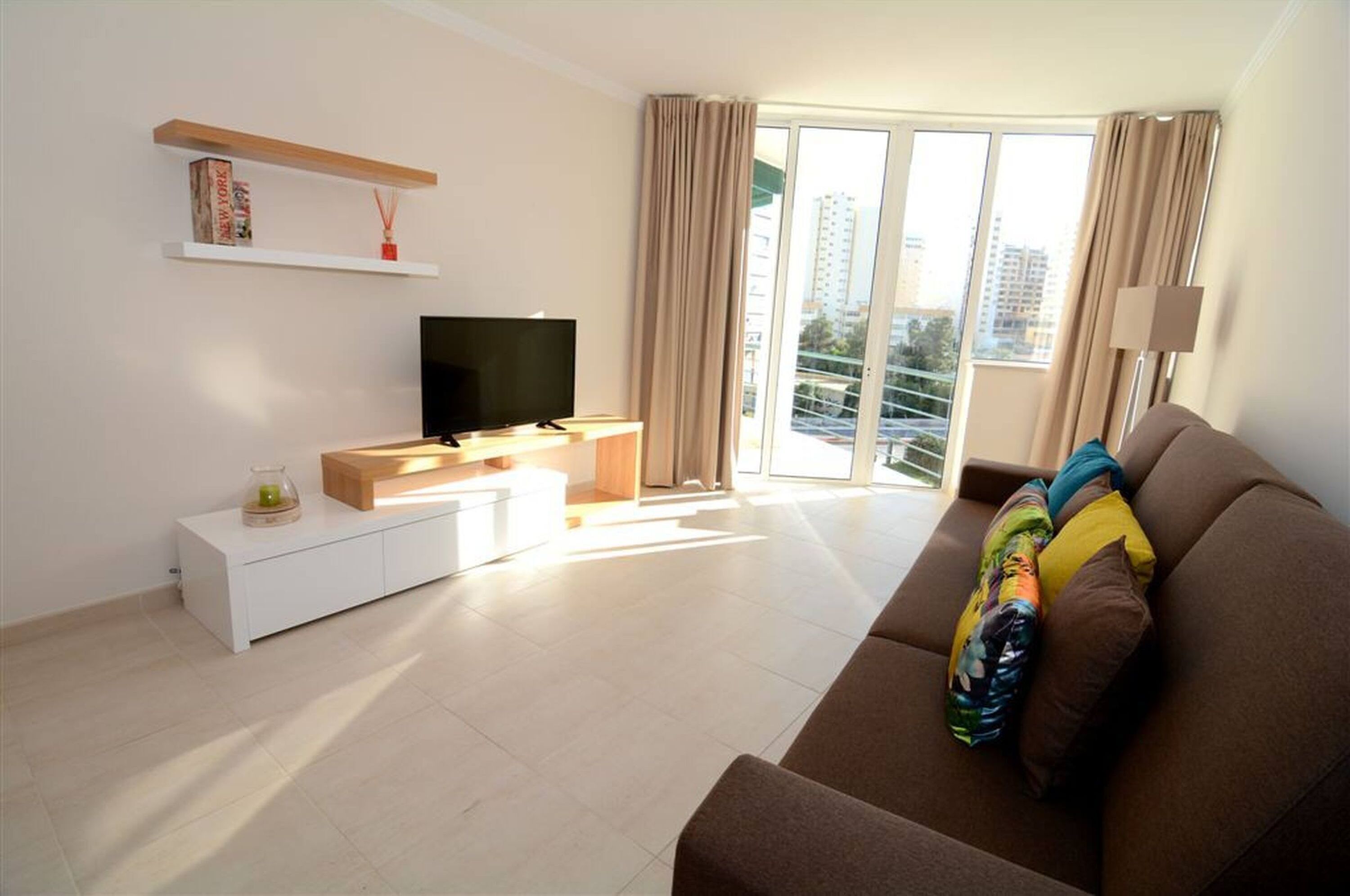 Ein modernes Wohnzimmer mit Flachbildfernseher, einem weißen Konsolentisch, einem braunen Sofa und einem Balkon mit Vorhängen.