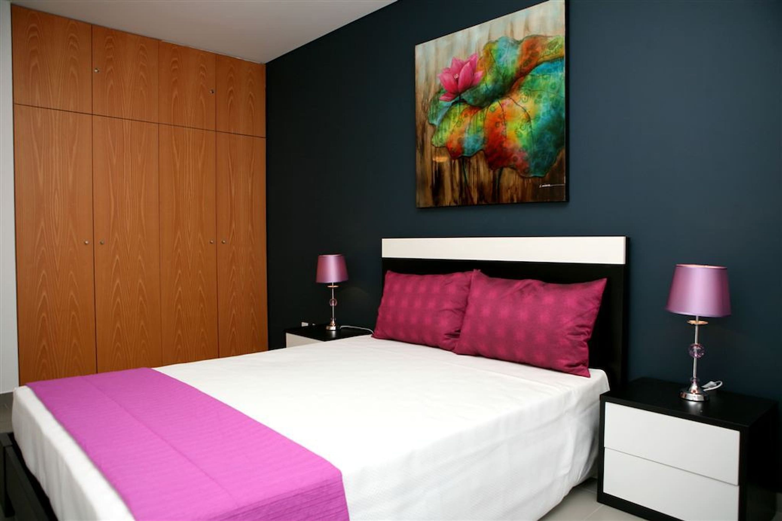 A bedroom with a bed, two bedside tables, a nightstand, a wooden wardrobe, and a colorful painting on the wall.