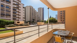 Un balcón con mesa y sillas de vidrio, vista a edificios altos y un área pavimentada con vegetación.
