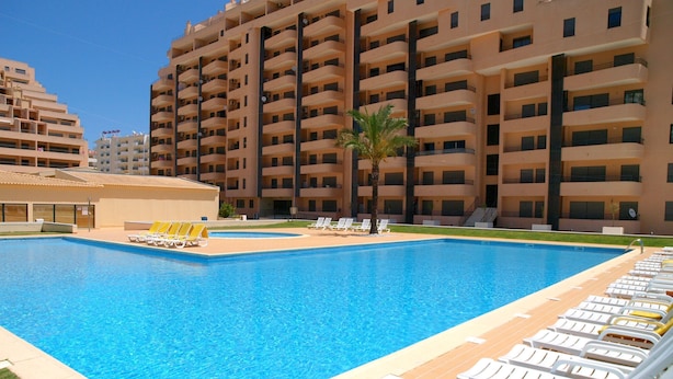 Una piscina con sillas de descanso y una palmera, rodeada por un edificio de varios pisos.