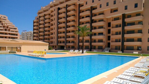 Una piscina con sillas de descanso y un edificio con varios balcones al fondo.