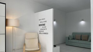 Ein modernes Wohnzimmer mit einem beige farbenen Sessel, einer Stehlampe und einem grünen Sofa. Eine weiße Wand zeigt ein Willkommen-Schild in verschiedenen Sprachen.