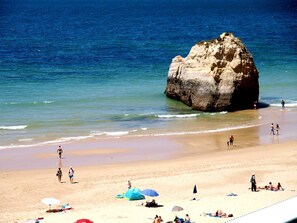 A beach with a large rock formation, clear blue water, and people enjoying the beach.