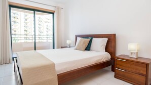 A modern bedroom with a wooden bed, a nightstand with a lamp, and a view of a residential building through a large window. - 27 - A420 in Portim o (Portimão)