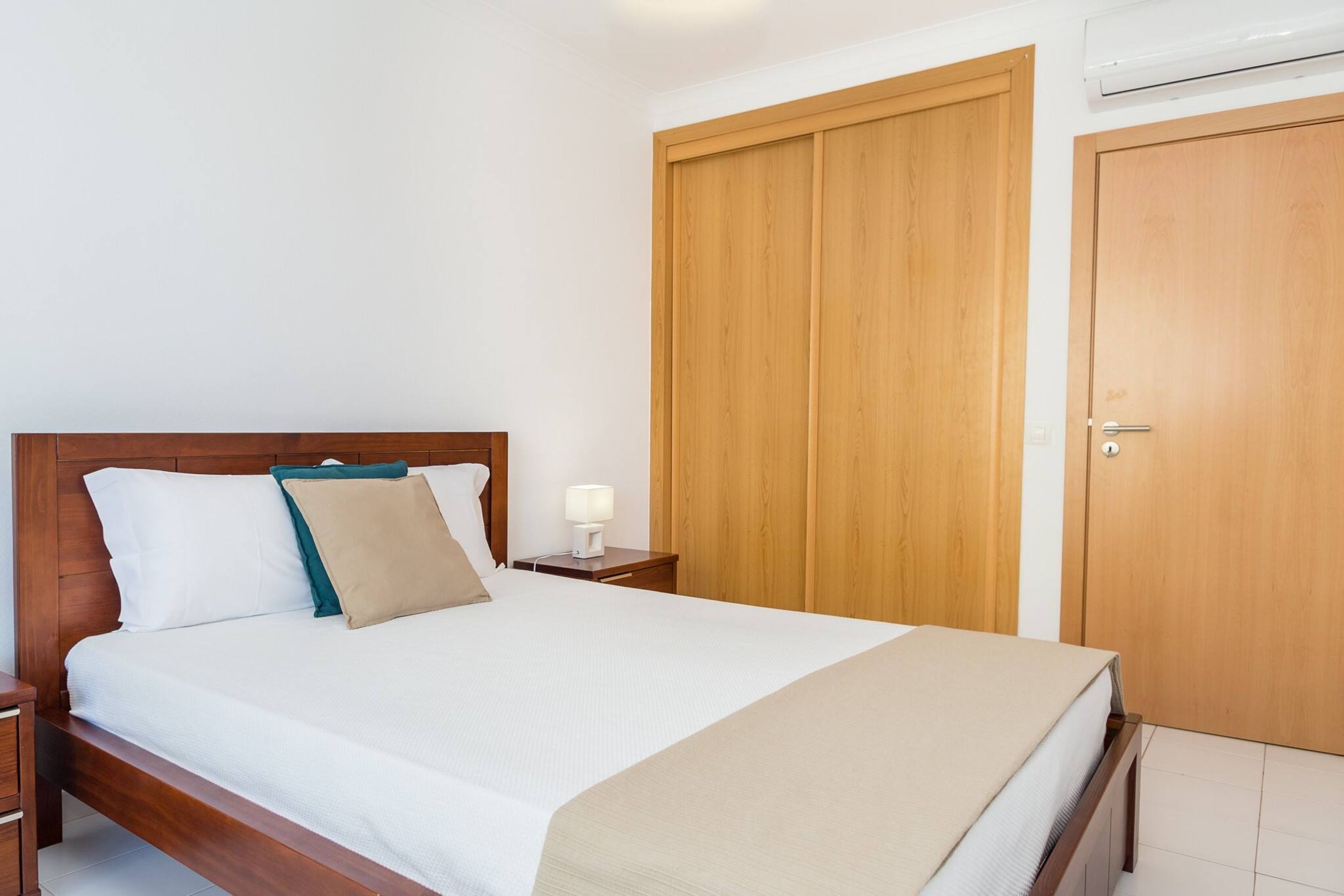 A neatly made bed with white linens and a beige throw, a wooden nightstand with a lamp, and a wooden wardrobe with a door.