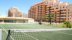 Un court de tennis avec un filet au premier plan, entouré d’immeubles résidentiels à plusieurs étages.
