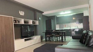 Un salon moderne avec un téléviseur à écran plat, un canapé, une table à manger avec des chaises, et un coin cuisine à l’arrière-plan.