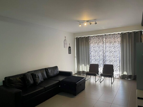 A modern living room with a black leather sofa, two armchairs, a coffee table, and a large window with patterned curtains.