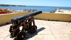 Un canon est installé sur une plateforme en pierre, surplombant une plage avec des bâtiments et un ciel dégagé.
