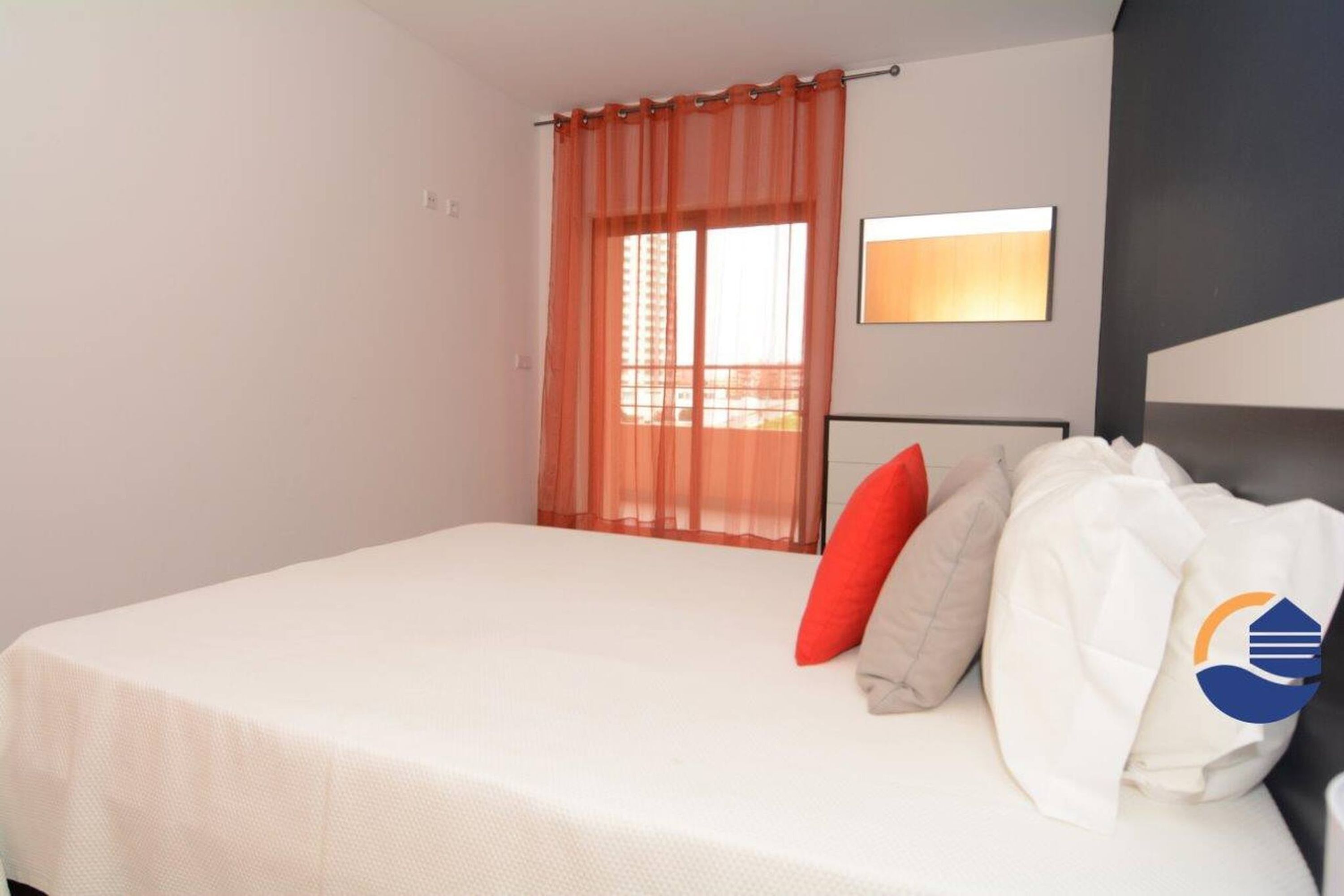 Een hotelkamer met een bed, een raam met oranje gordijnen, een nachtkastje en een spiegel aan de muur.