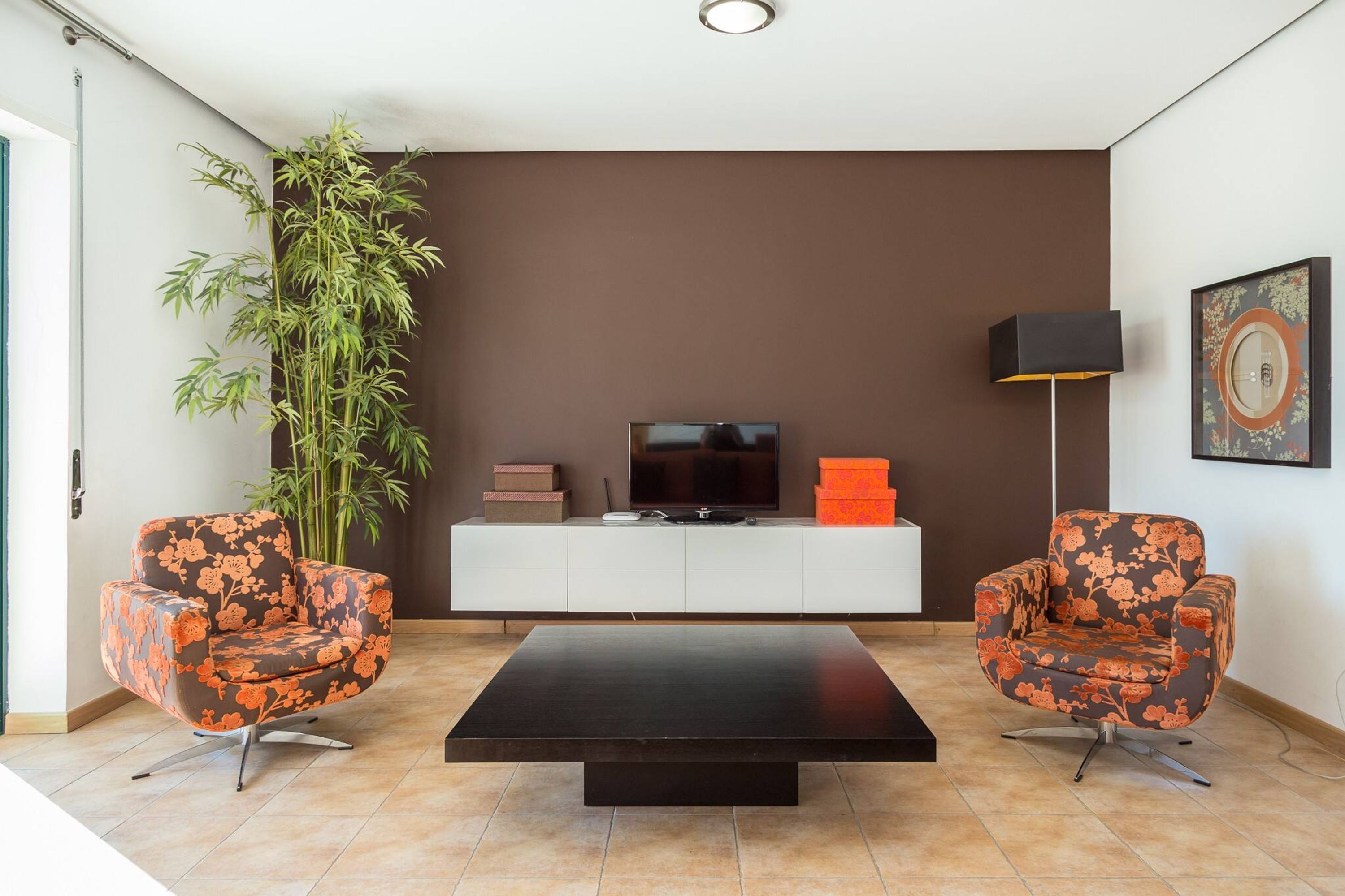 Una sala moderna con una mesa de centro negra, dos sillones con motivos florales, una planta grande en maceta, un televisor sobre un mueble blanco y una lámpara de pie.