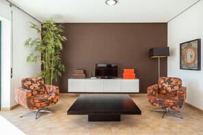 Ett modernt vardagsrum med ett svart soffbord, två blommiga fåtöljer, en stor krukväxt, en tv på ett vitt skåp och en golvlampa.