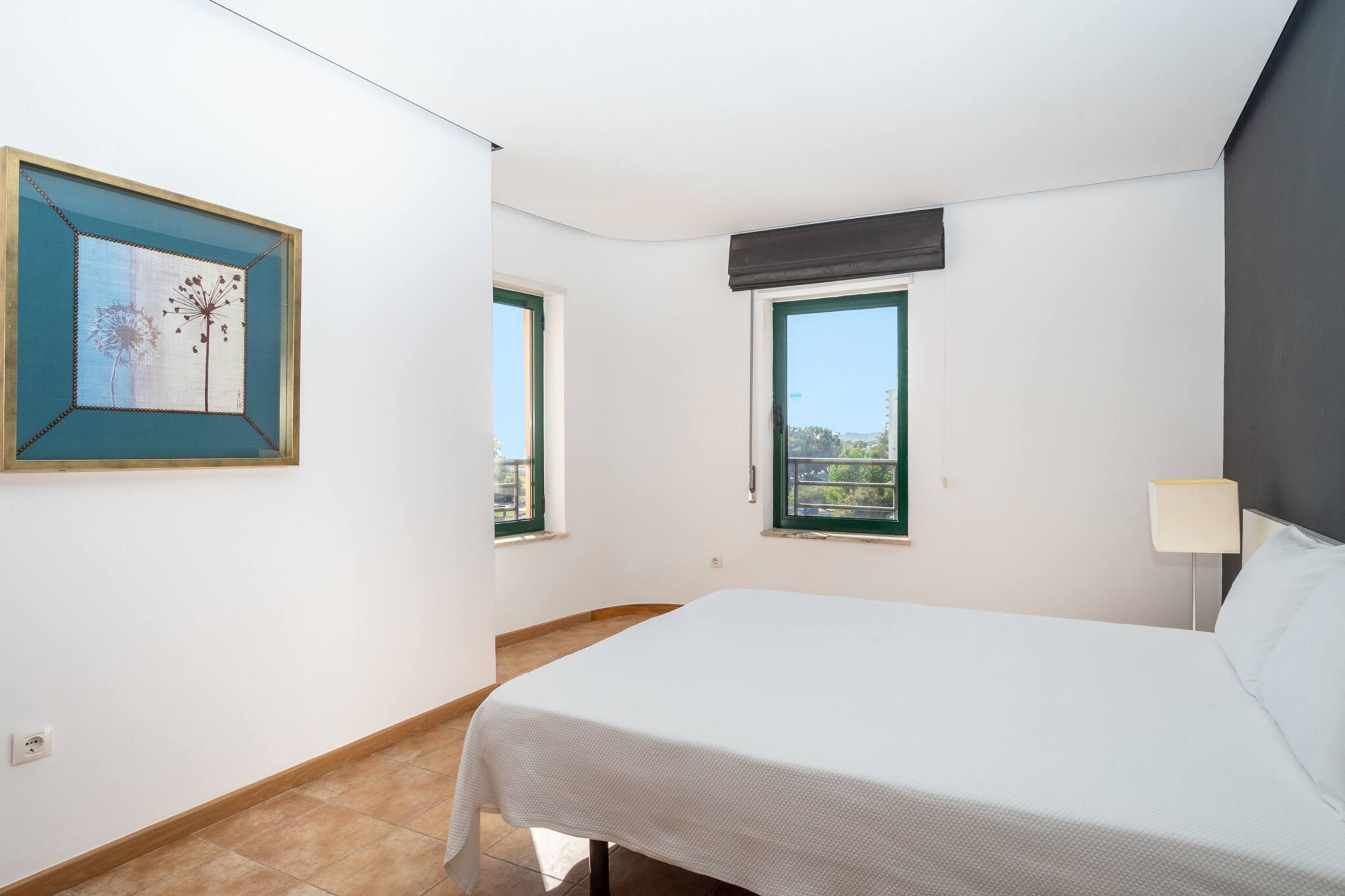 A bedroom with a bed, a window, a wall-mounted lamp, and a framed picture.