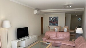 A modern living room with a flat-screen TV, a glass coffee table, a pink sofa, and a dining area with chairs and a table. - 41 (Portimão)
