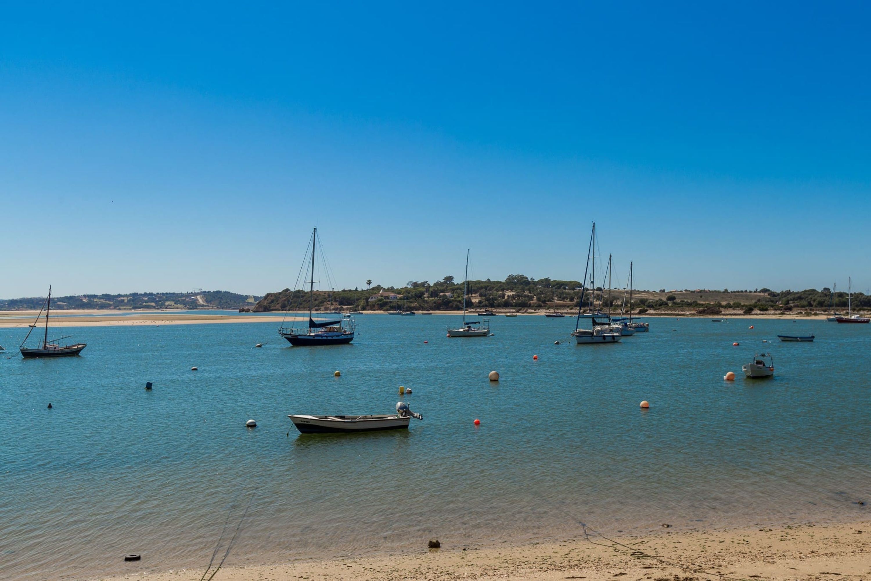 A coastal scene with boats anchored in calm waters, a sandy beach, and a distant shoreline with buildings.