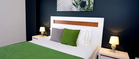 Una cama bien hecha con una colcha verde, almohadas blancas y un cabecero de madera. Hay dos mesitas de noche con lámparas, y una obra de arte colgada en la pared sobre la cama.