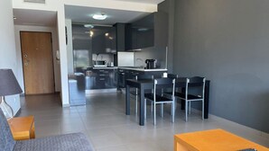 A modern living room with a grey sofa, a wooden coffee table, and a TV. A kitchen area with dark cabinets and a dining table with chairs is visible in the background. - 13 - B315 in Portim o (Portimão)
