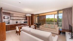 Un salon moderne avec un canapé, un coin repas avec une table et des chaises, une cuisine équipée et un balcon offrant une vue sur la verdure.