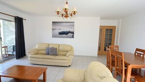 Ett vardagsrum med en beige soffa, ett soffbord i trä, ett matbord med stolar och en kristallkrona.