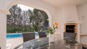 A glass-top table with wicker chairs, a white vase with flowers, and a fireplace in a room with a view of a swimming pool and trees.