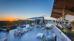 A rooftop terrace with white furniture, a covered seating area, and a view of the sunset.