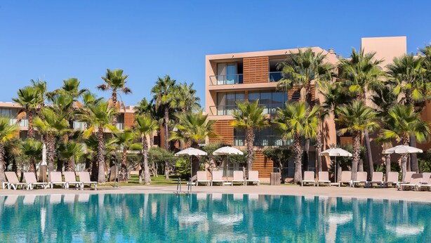 A poolside area with lounge chairs and umbrellas, surrounded by palm trees and modern buildings.