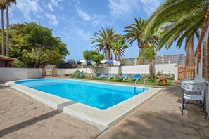 A rectangular swimming pool with clear blue water, surrounded by lounge chairs and umbrellas, palm trees, and a stone patio.