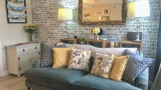Una sala de estar con un sofá azul, almohadones decorativos, una mesita de madera y un espejo fijado a la pared.