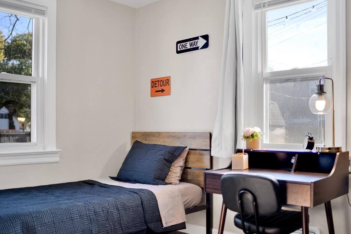 Une chambre à coucher avec un lit, un bureau et une chaise. Il y a deux fenêtres et une porte.