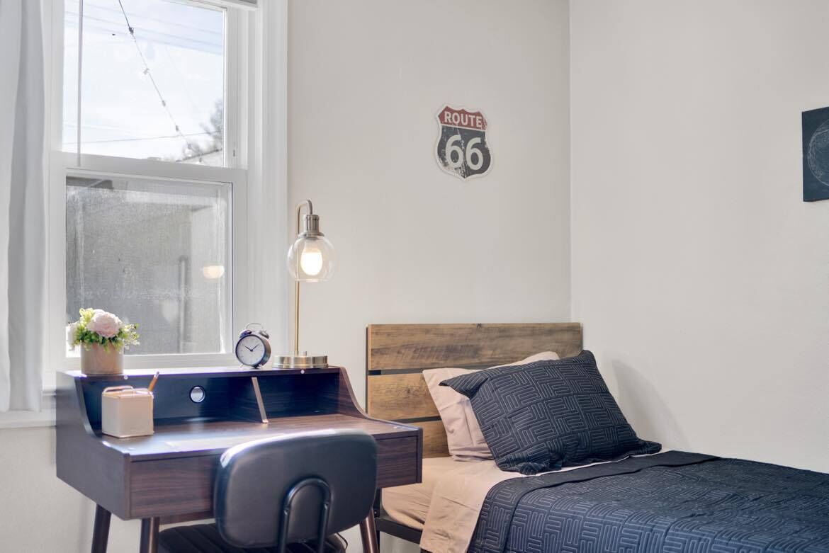 Ein Schlafzimmer mit hölzernem Kopfteil, einem Bett mit gemusterten Betttextilien, einem Schreibtisch mit Lampe, einer Uhr und einem Route 66 Schild an der Wand.