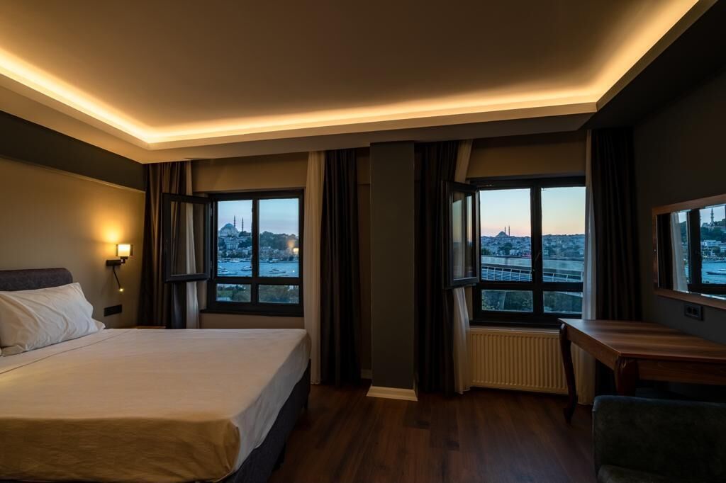 A hotel room with a large bed, a desk, and a view of a cityscape through the windows.