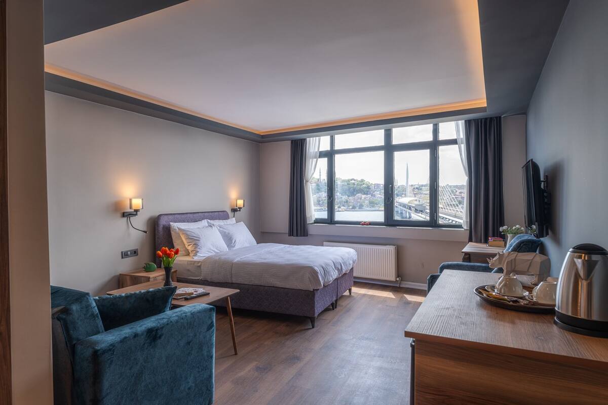 A modern hotel room with a large bed, a desk with a kettle, a chair, and a view of the city through large windows.