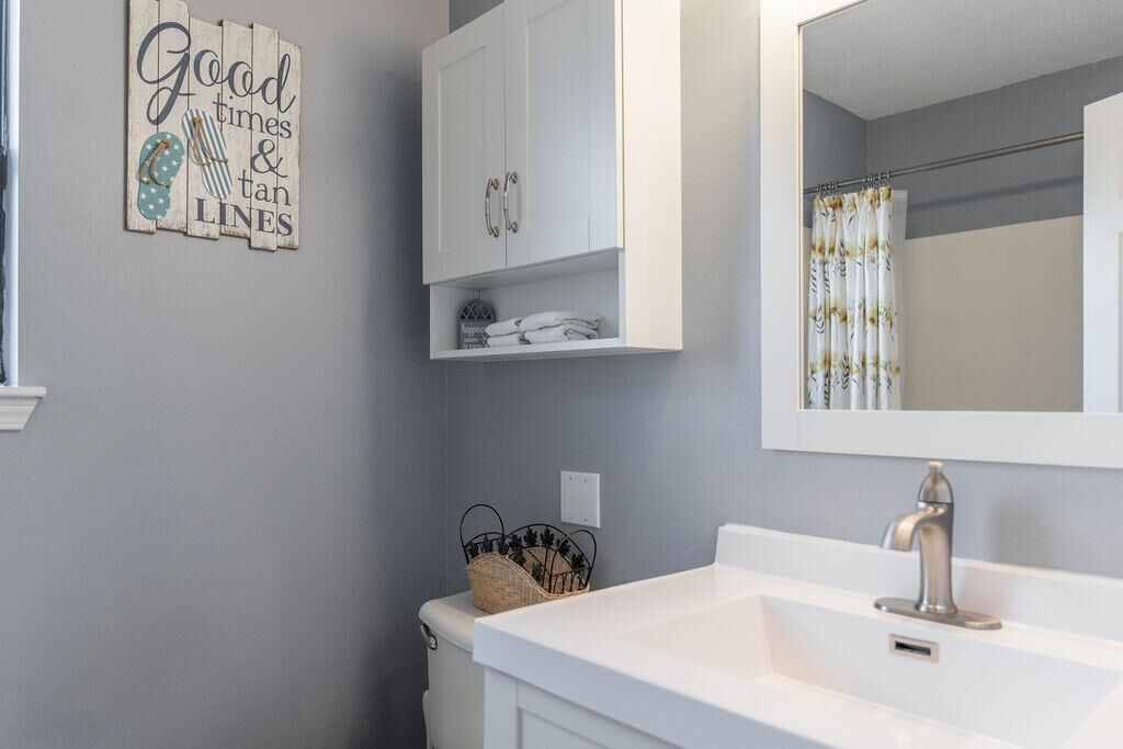 Un baño con un lavamanos blanco, un espejo y un perchero para toallas. Hay una decoración de pared con la inscripción 'Good times & tan lines' y una cesta en el suelo.