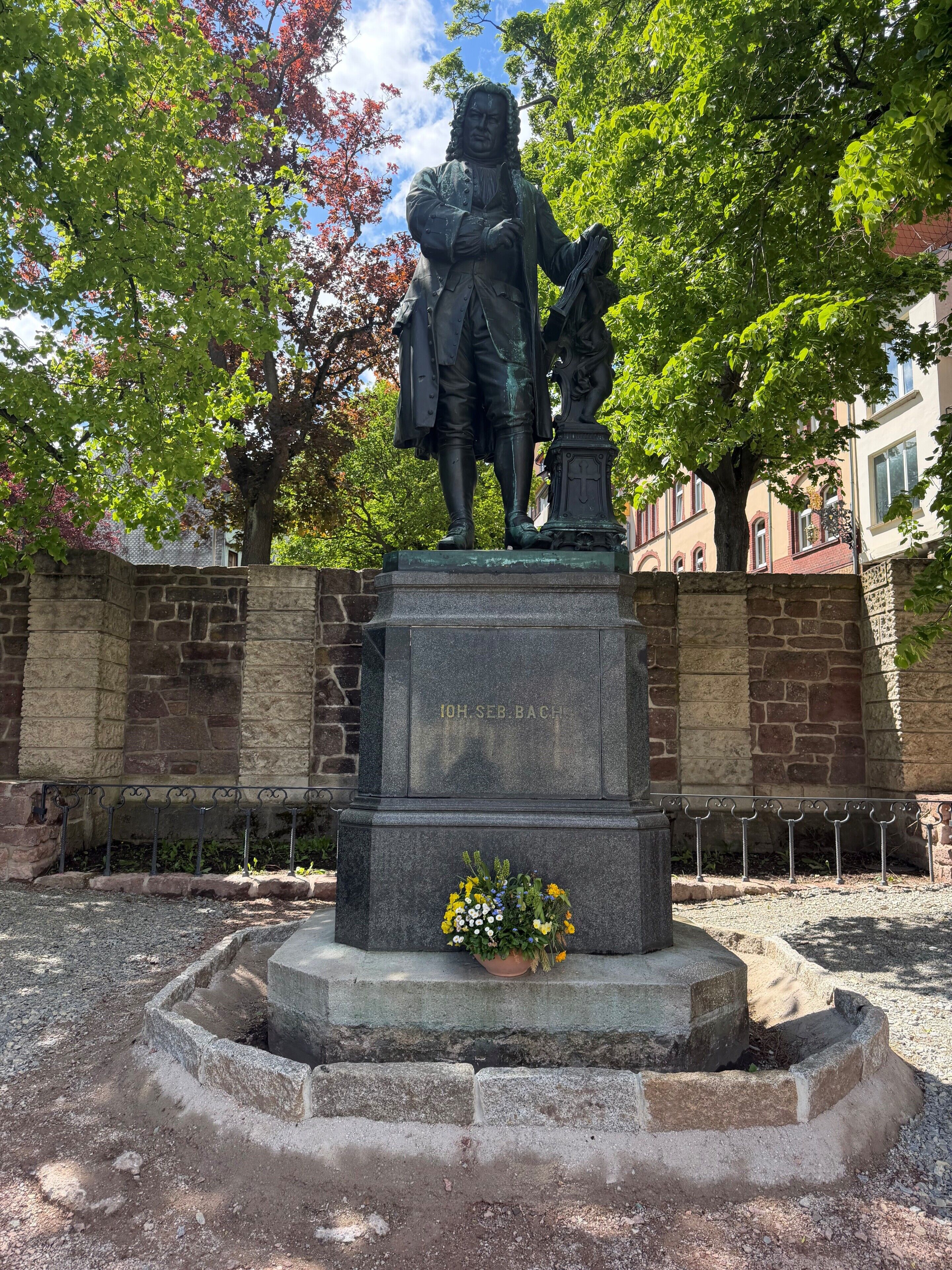 A statue with a pedestal, surrounded by a stone wall and trees.