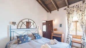 Une chambre à coucher avec un lit, des oreillers, une chaise en bois, une petite table et une fenêtre avec des rideaux.