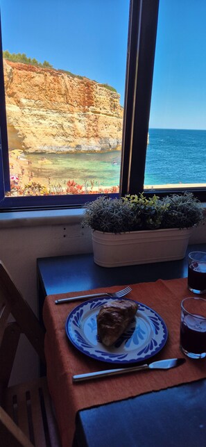 A dining table with a plate of food, a glass of drink, and a window view of a beach and cliffs. - Bungalow on Benagil Beach (Benagil)