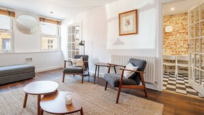 En moderne stue med en grå sofa, to lænestole, et sofabord og en gulvlampe. Der er et stort spejl på væggen, et billedramme og en radiator.